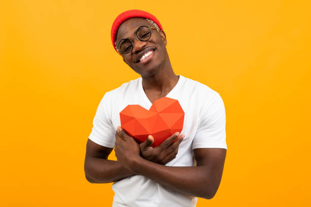 cute handsome american in white t-shirt holds 3d heart made of paper for valentines day on yellow background.